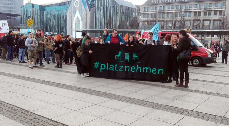 Mehrere Demonstrierende am Leipziger Augustusplatz mit einem Banner vom Aktionsnetzwerk Leipzig nimmt Platz.