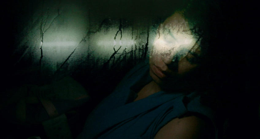 Eine junge Frau schläft in einem Bus, ihr Kopf lehnt am Fenster. Sie trägt ein blaues Oberteil. Es ist sehr dunkel, das einzige Licht der Neonröhren verschwimmt in der beschlagenen Scheibe.
