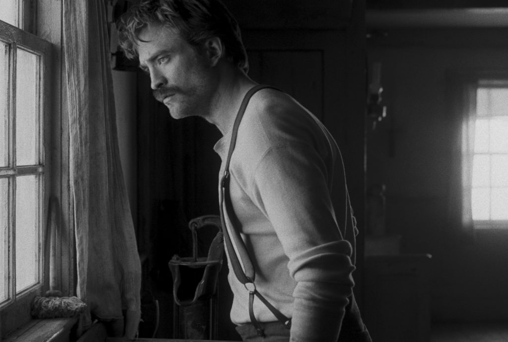 Robert Pattinson alias Ephraim Winslow schaut besorgt aus dem Fenster des Wärterhäuschens. Er trägt eine Hose mit Hosenträgern und einen für die 1900er Jahre typischen Schnauzer.