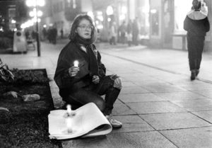 Alternativtext: Eine junge Frau mit erschrockenem Ausdruck sitzt in einer Fußgängerzone auf dem Boden. Sie hält eine Kerze in der Hand, neben ihr stehen zwei weitere, im Hintergrund Passanten.
