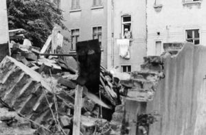 : Im Vordergrund ein Haufen von Trümmern und Schutt, ein Stück Mauer steht noch. Dahinter hängt eine Frau auf der Leine vor ihrem Fenster Wäsche auf.