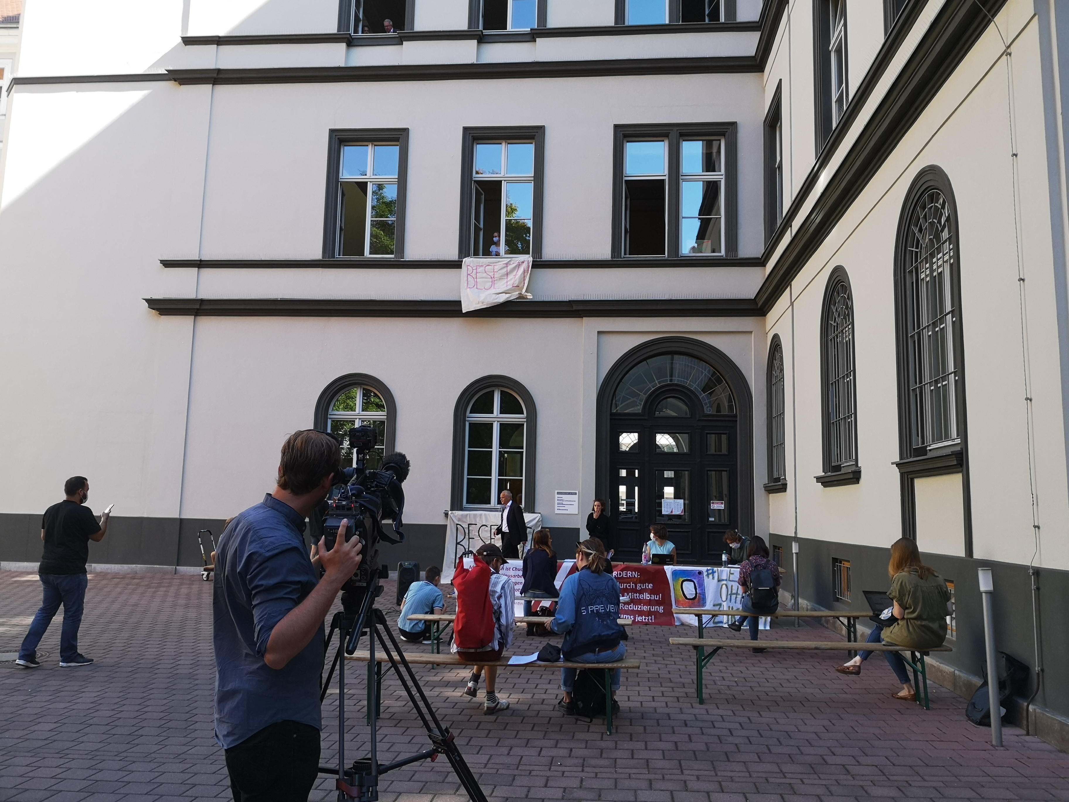 Der Innenhof des Rektorats in der Ritterstraße, im Vordergrund eine Fernsehkamera, im Mittelgrund Bänke, auf denen Journalist*innen sitzen, im Hintergrund das Rektorat, aus dem ein Banner mit der Aufschrift "besetzt" hängt.