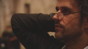 Christoph Schlingensief ist im Profil vor einem dunklen Hintergrund zu sehen. Sein Gesicht ist mit warmem Licht beleuchtet und er blickt aufmerksam auf einen Punkt hinter dem linken Bildrahmen. 