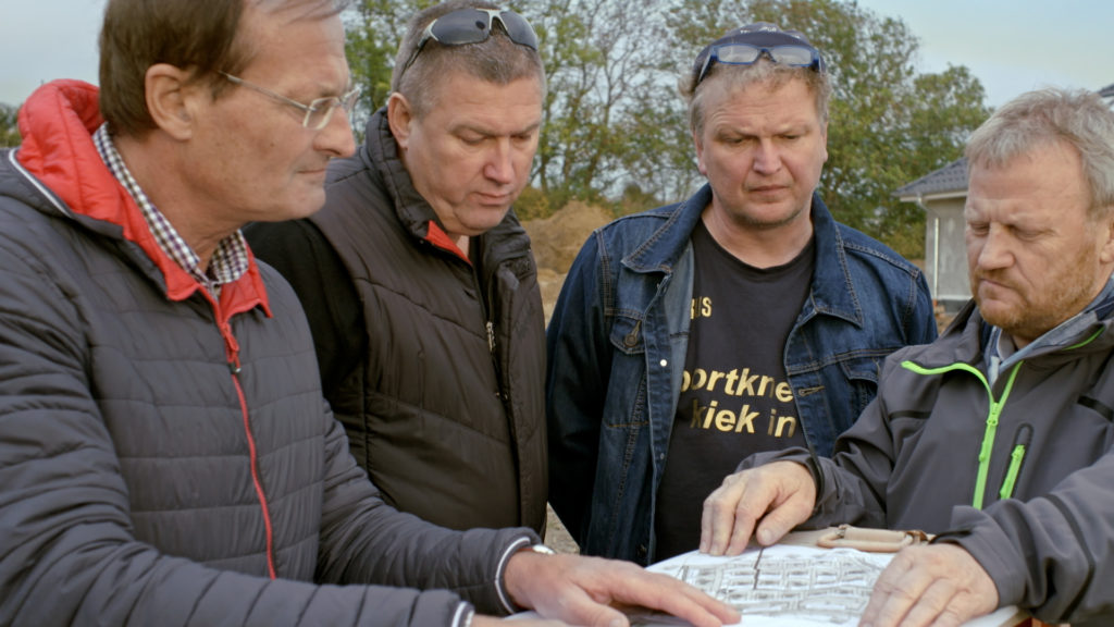 Edwin, Klaus, Markus und Hans-Harald stehen nebeneinander, halten eine Karte in den Hän-den und blicken darauf. Sie sind die „Vier von der Stange“ und planen den Bau des Neubau-gebiets.