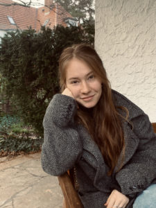Hannah sitzt draußen auf einer Terasse und lächelt in die Kamera. Sie trägt eine schwarz-weiße Jacke. Im Hintergrund sieht man die Hauswand und eine Hecke.