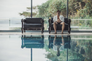 David sitzt in einem Ledersessel an einem Indoor-Pool, an seiner Seite ein Glas Orangensaft. Er trägt Badeshorts, Flipflops und ein offenes weißes Hemd.