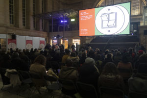 Großes Publikum in der Bahnhofshalle. Auf dem Bildschirm Dok Logo mit zwei Lichtschaltern.