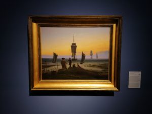 Ein Gemälde hängt an einer blauen Wand. Auf dem Gemälde sind evrschieden alte Menschen am meer zu sehen. Im Hintergund Segelschiffe.
