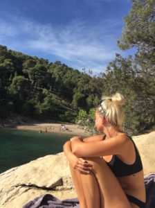 Kolumnistin Leoni sitzt in einem schwarzen Bikini auf einem Felsen, die Arme um die angezogenen Knie geschlungen, und schaut auf das Meer, den Strand und den umgebenden Wald.