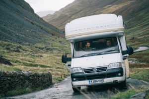  Sam und Tusker fahren mit einem weißen Wohnwagen durch Nordengland. Sie fahren eine kurvige Straße entlang, die sich zwischen ein paar Bergen hindurch schlängelt.