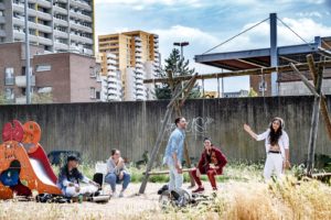 Die migrantischen Jugendlichen sitzen auf einem Spielplatz und spielen Werwolf. Naima zeigt die Werwolfkarte, die anderen schauen überrascht und verärgert. Im Hintergrund sind hohe Plattenbauten zu sehen.