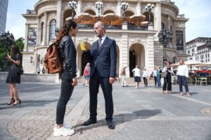 Naima und Pohl stehen vor der Alten Oper in Frankfurt am Main. Er drückt ihr ein dünnes, gelbes Reclam-Heft in die Hand. Im Hintergrund stehen Passanten.