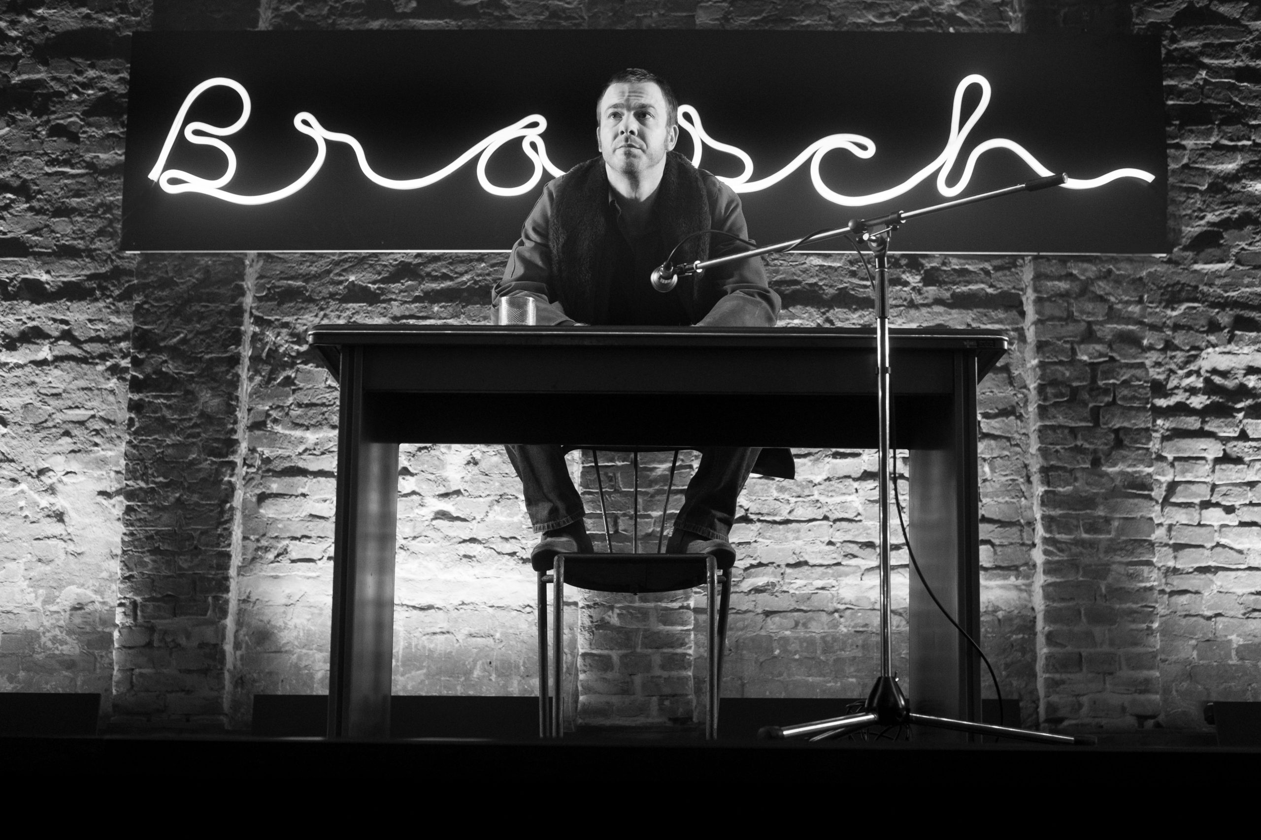 Brasch sitzt auf der Lehne eines Stuhls vor einem Tisch; hinter ihm leuchtet der Neonschriftzug „Brasch“.