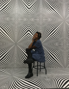 Funmi Olanigan sitzt auf einem Stuhl vor einem prisma-mäßig verzerrten Hintergrund.