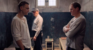Hans und Viktor stehen vor ihren Betten in der Zelle, während ein Wärter die Zelle durchsucht.