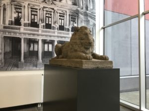 Der steinerne Löwe liegt auf einem Sockel, der wiederum auf einem ein meter hoen Quader steht. Im Hintergrund hängt ein Foto, welches die Uni leipzig vor ungefähr hundert Jahren zeigt.