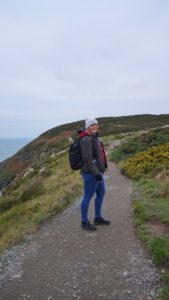 Natalie Stolle trägt eine dicke Jacke, eine Mütze und einen Rucksack. Sie schaut in die Kamera und steht auf einem Wanderweg in felsiger Landschaft. Die Vegetation ist sehr niedrig und scheint einem stetigen Wind trotzden zu müssen.