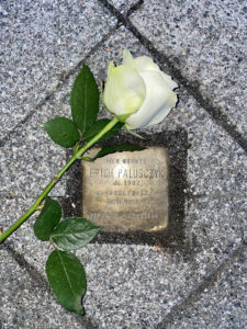 Das Foto zeigt einen kupfernen Stolperstein eingelassen in eine Gewegplatte. Eine weiße Rose mit grünen Blättern liegt am Stein. Auf dem Stein steht: "Hier wohnte Erich Palusczyk, Jg. 1902, ermordet 1940, (dann folgen zwei Zeilen, die ich nicht lesen kann)".