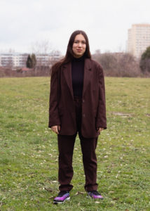 Ronya Othmann steht auf einer Wiese in einem großen braunen Anzug.