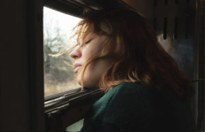 Eine rothaarige Frau, die Protagonistin Laura, hat ihren Kopf an dem offenen Zugfenster angelegt, ihre Haare wehen im Wind, ihre Augen sind geschlossen.