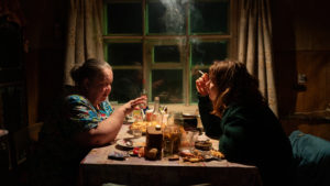 Zwei Frauen sitzen an einem Tisch, draußen ist es dunkel. Die Frau auf der linken Seite ist schon alt, sie hat graue Haare. Die Frau auf der rechten Seite, Laura, ist im Studentenalter und raucht eine Zigarette. Auf dem Tisch stehen Leckereien und die beiden unterhalten sich.