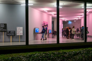 Man befindet sich außerhalb der Ausstellungsräume des GfZK und schaut durch die Glasfassade hinein. Rechts sieht man Leute, welche sich die AUstellung anschauen. Links lehnen Schilder an der Wand, sie gehören auch zur Ausstellung. Auf ihnen steht zum Beispiel: Nie wieder Faschismus!; Kein Schlussstrich