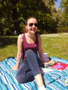 Das Bild zeigt Kolumnistin Sophie auf einer gestreiften Decke im Park sitzend. Sie trägt ein rotes Top, eine graue Jeans und hat eine vor der hellen Sonne schützende Sonnenbrille auf, während sie mit einem Lächeln in Richtung der Kamera schaut. Hinter ihr ist eine blühende Wiese und ein großer, schattenspendender Baum zu sehen.