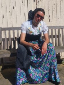 Rahel Selnack – ein braunhariges Mädchen anfang 20 – auf grauer Bank und mit Zigarette in der Hand in ihrer Tracht. Diese besteht aus einem weißen Oberteil mit gelben gestickten Blumen und einem lila, blau, weiß und grün geblümten langen Rock, sowie einer schwarzen Schürze, schwarzer Kopfbedeckung.