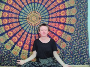 Kolumnistin Naomi sitzt vor ihrem Mandala-Wandbehang und meditiert.