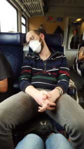 Kolumnist Franz sitzt auf einem Sitzplatz in einem Zug der österreichischen Bundesbahn. Er trägt eine beige Hose, einen buten Pulli, eine FFP2-Maske und schläft.
