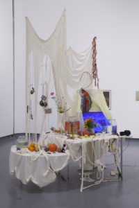 Der Altar des Kollektivs Polymora Inc. ist der Yorubà-Göttin Oshun geweiht. Er ist bedeckt mit weißen Tüchern. Auf ihm befinden sich Obst, Gemüse, mehrere Behälter mit Wasser, Fotos, Blumen und ein Bildschirm, der die Videoinstallation des Kollektivs zeigt.