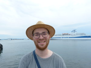 Kolumnist Franz lächelt in die Kamera, im Hintergrund sind das Meer und ein Schiff zu sehen