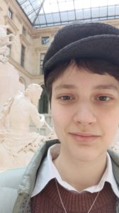 Ein Porträt von Kolumnistin Laura, die am rechten Bildrand direkt in die Kamera lächelt. Schwarze Schiebermütze, unter der kurze, braune haare hervorgucken, braune Augen eine kakifarbene Jacke, darunter ein brauner Strickpulli unter dem der Kragen eines weißen Hemdes hervorschaut. Am linken Bildrand im Hintergrund eine weiße Mamorstatue mit langen gelockten haaren und ein sandsteinfarbenes Gebäude mit pyramidenförmigen Glasdach.