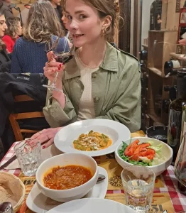 Kolumnistin Lene sitzt mit einem Glas Wein in der Hand vor einem gedeckten Tisch in einem Restaurant; im Hintergrund sind Menschen zu sehen