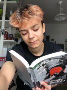 Leen hält das Buch, im Hintergrund eine Schrankwand.