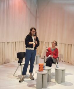 Lisa Weeda mit der Übersetzerin Birgit Erdmann. Weeda steht, Erdmann schaut sie an. Vor ihnen ist Weedas neuer Roman aufgestellt.