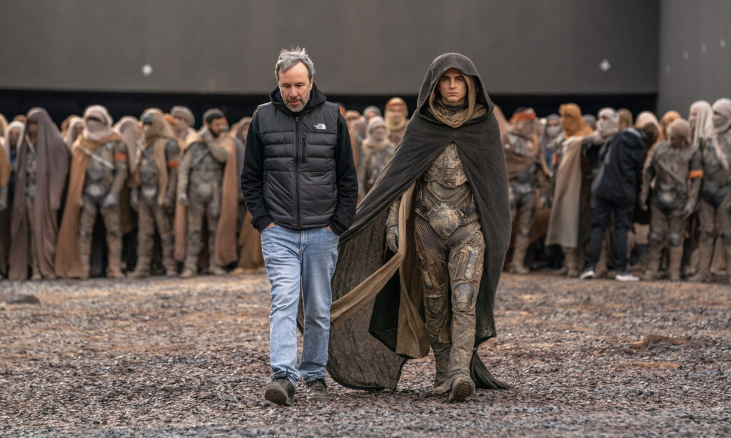 Im Bild sieht man den Regisseur Denis Villeneuve mit dem Schauspieler Timothée Chalamet. Sie gehen auf die Kamera zu. Der Regisseur schaut auf den Boden, Chalamet nach vorne. Letzterer trägt ein Kostüm aus dem Dune-Universum. Im Hintergrund ist eine Menge weiterer Schaupieler. 