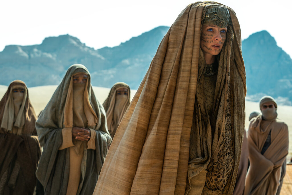 Man sieht die Schauspielerin Rebecca Ferguson in ihrer Rolle als Lady Jessica. Sie scheint vorwärts zu gehen und schaut nicht in die Kamera. Sie trägt die Kleidung der Fremen. Im Hintergrund befinden sich weitere Fremen. 